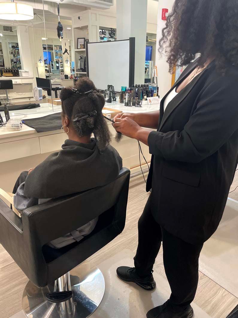 Hair stylist styling client's hair in a salon. Client is seated. Stylist wears black outfit.