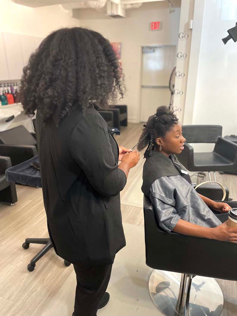 Hair stylist working on a client's hair in a salon; client is seated, wearing a protective cape.