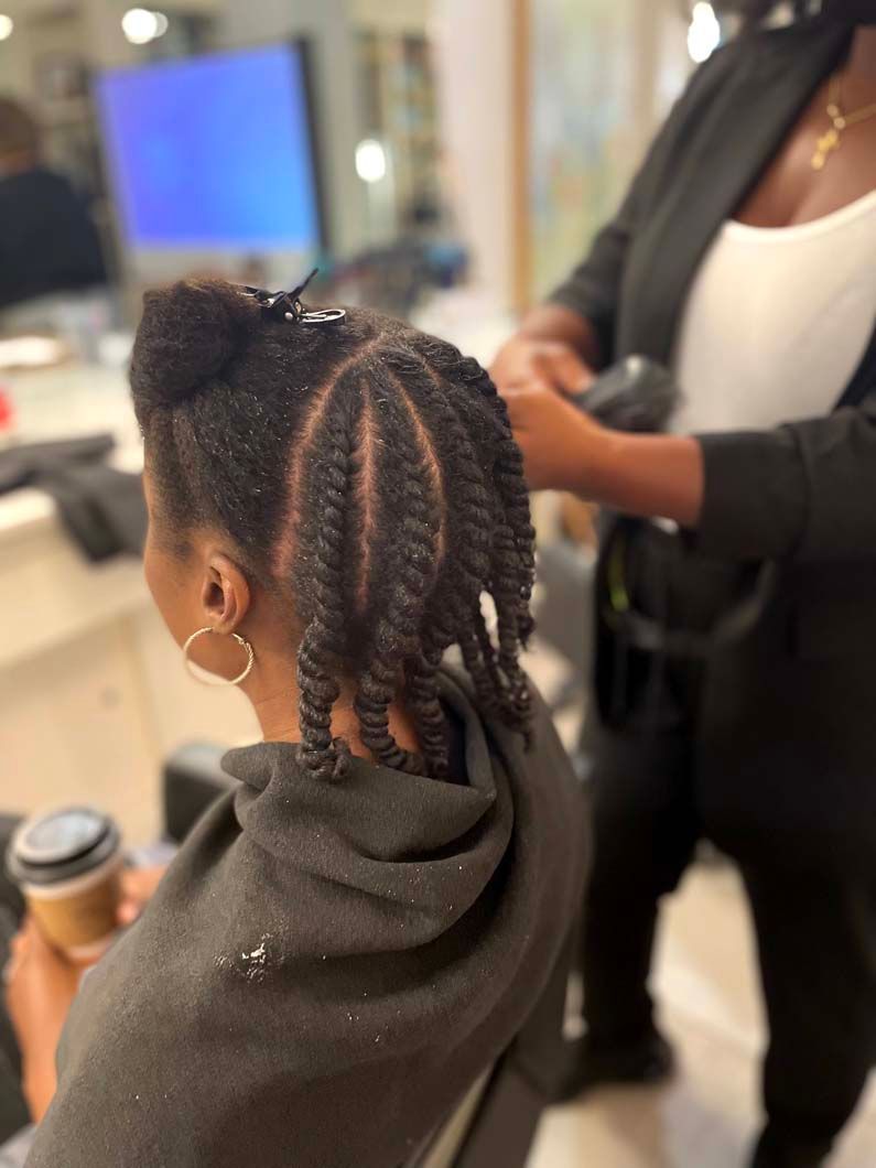 Person getting hair styled into twists at a salon, another person working on their hair.