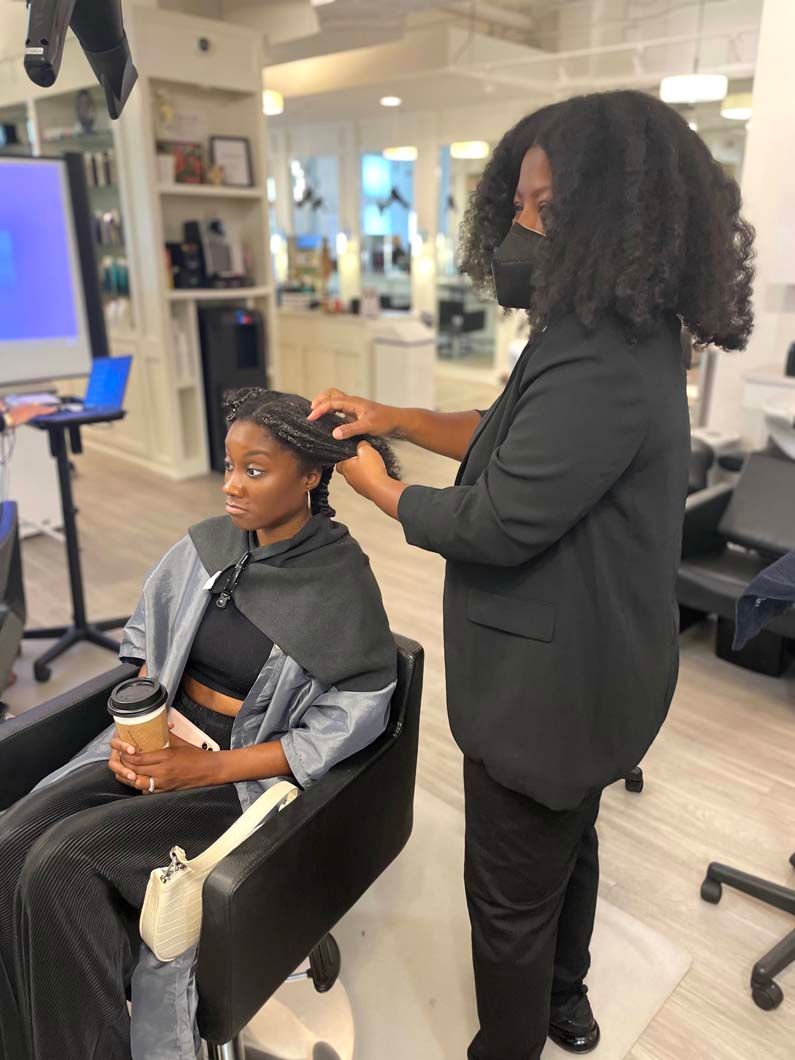 Hair stylist working on client's hair in a salon; client seated, wearing cape and holding coffee.