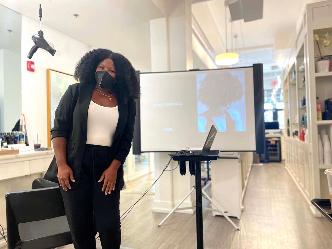 Woman wearing a mask and black suit stands by a presentation screen in a salon.