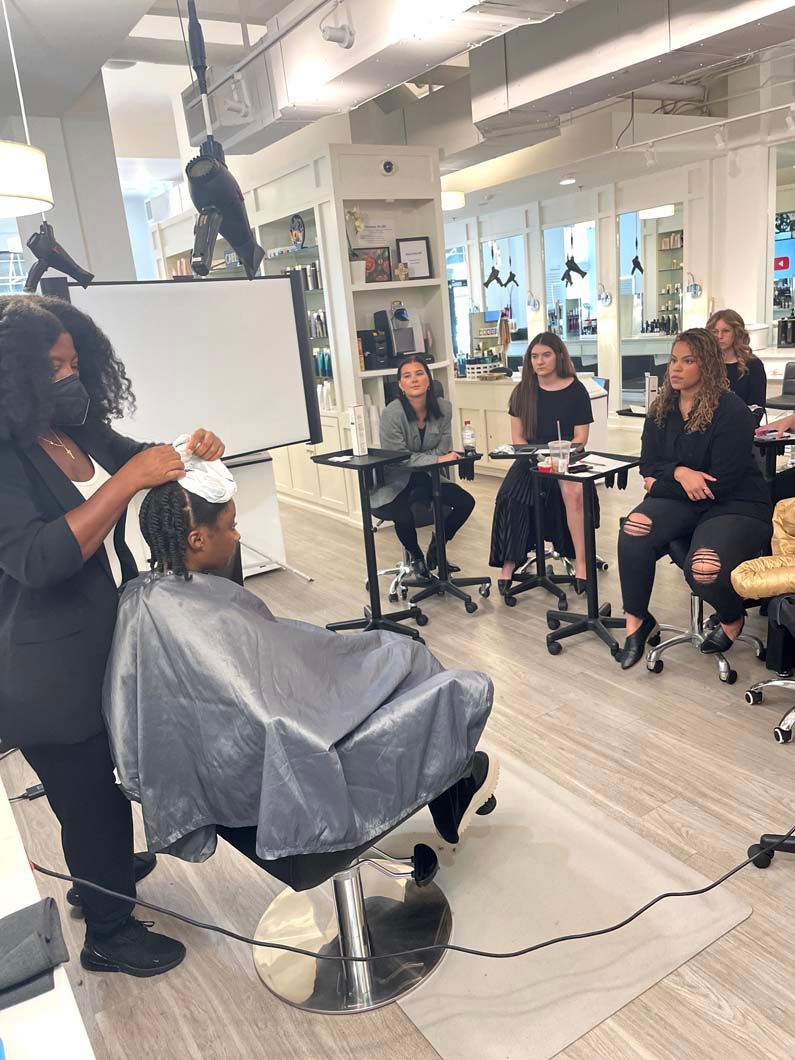 Hairdresser working on a client's hair during a demonstration in a salon setting, with an audience observing.