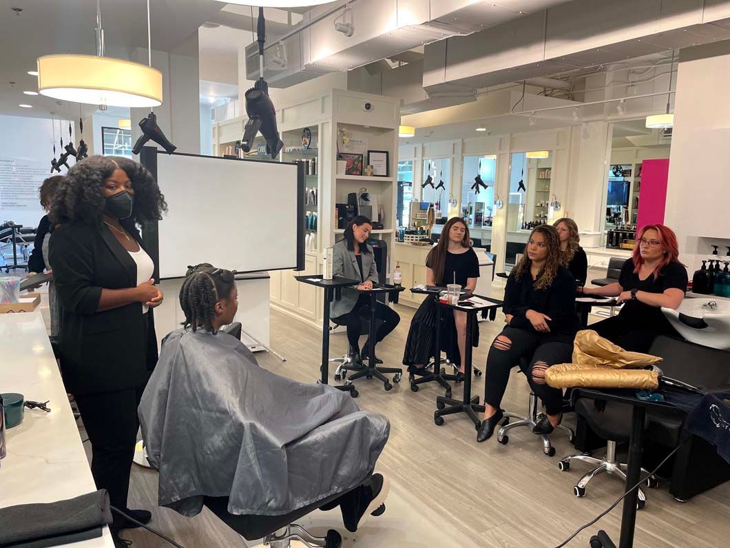 A hairstyling class in a salon. A stylist speaks to students watching a woman in a styling cape.