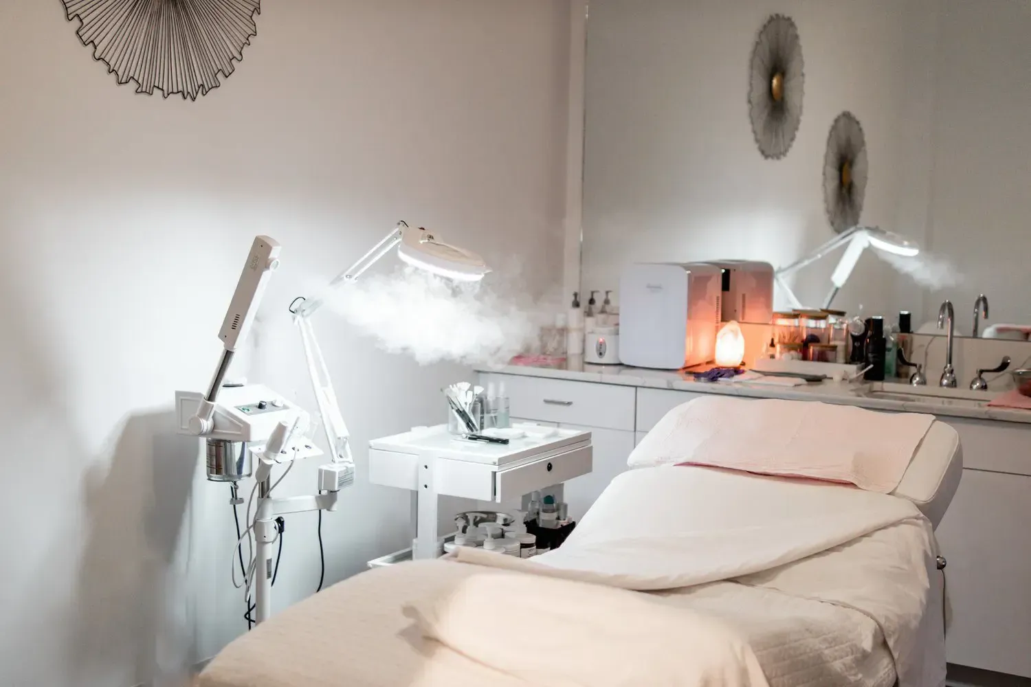 Facial treatment room with bed, equipment, and a misting machine, all in white.