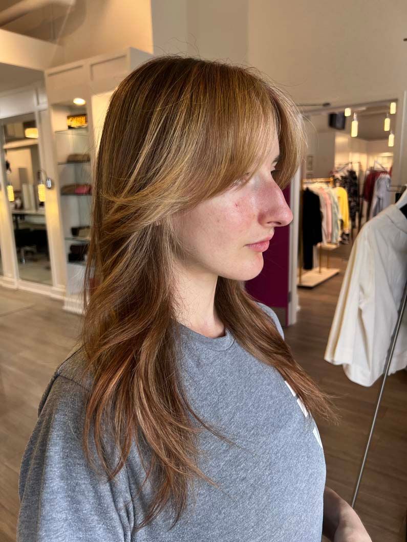 Woman with layered, reddish-brown hair and side-swept bangs indoors, near a clothing rack.