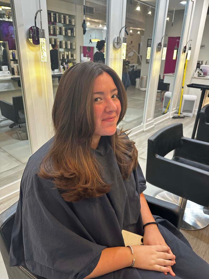 Person in a salon chair with layered brown hair, smiling.  Wearing a black cape. Well-lit salon setting.