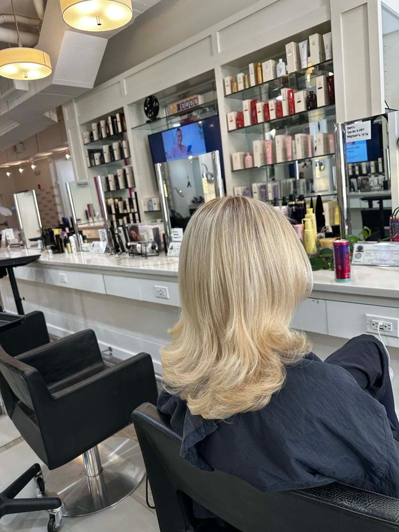 Woman with crimped blonde hair sits in a salon chair. Salon interior with products on shelves and mirrors.