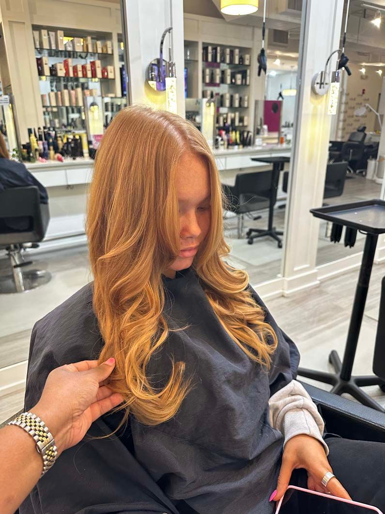 Woman with long, wavy, copper-colored hair in a salon. A hand adjusts her hair.