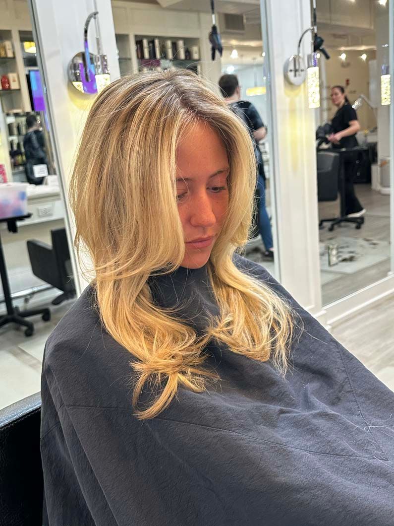 Woman with blonde crimped hair in a salon, wearing a salon cape, looking down.