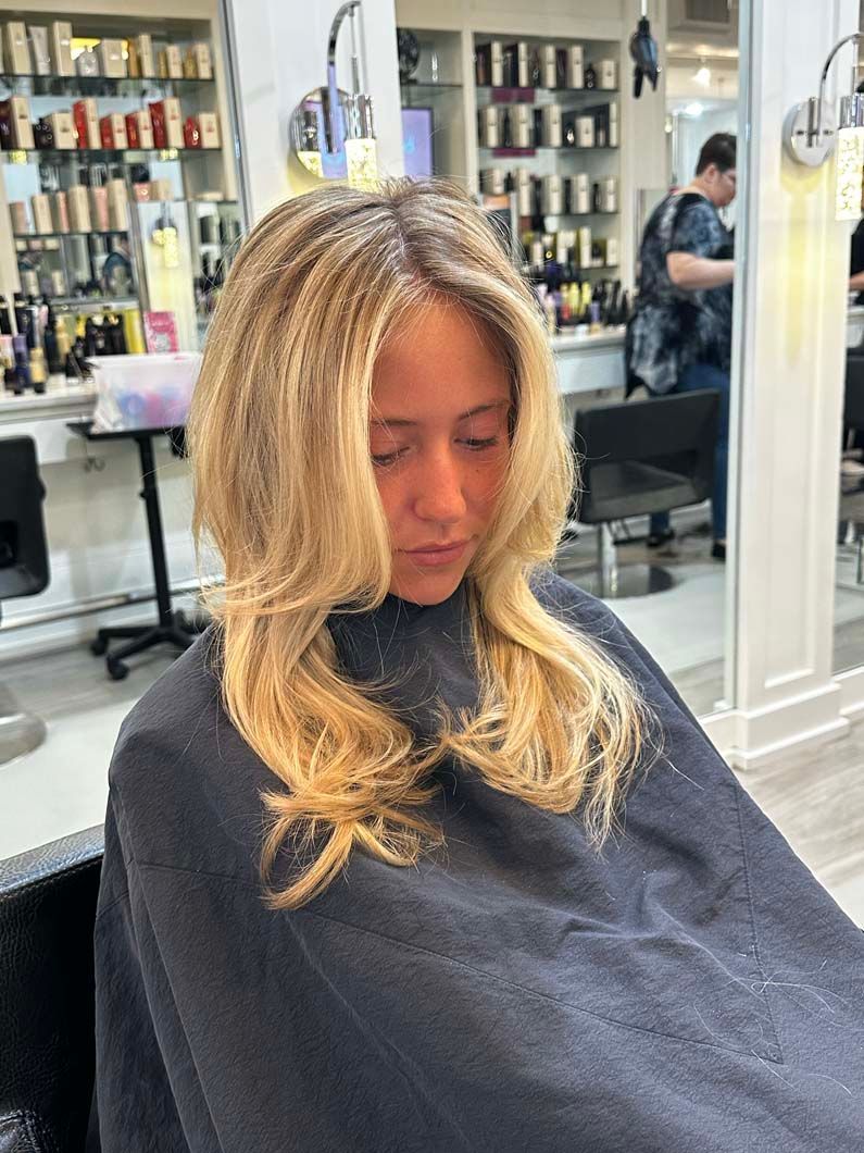 Woman with blonde hair in a salon chair, wearing a cape, having her hair styled.