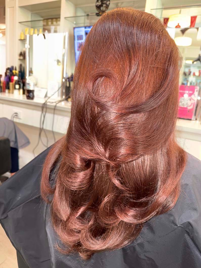 Woman with reddish-brown hair styled in waves and curls, seated in a salon chair.