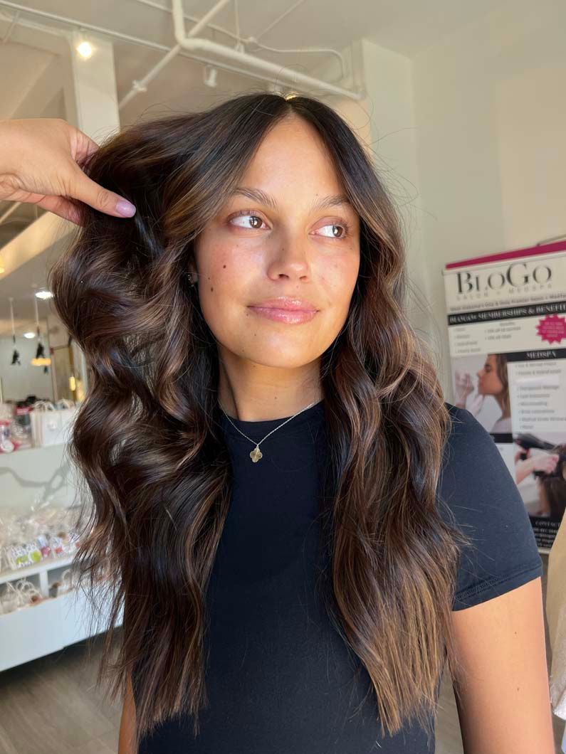 Woman with long wavy brown hair, wearing a black shirt, in a salon setting.