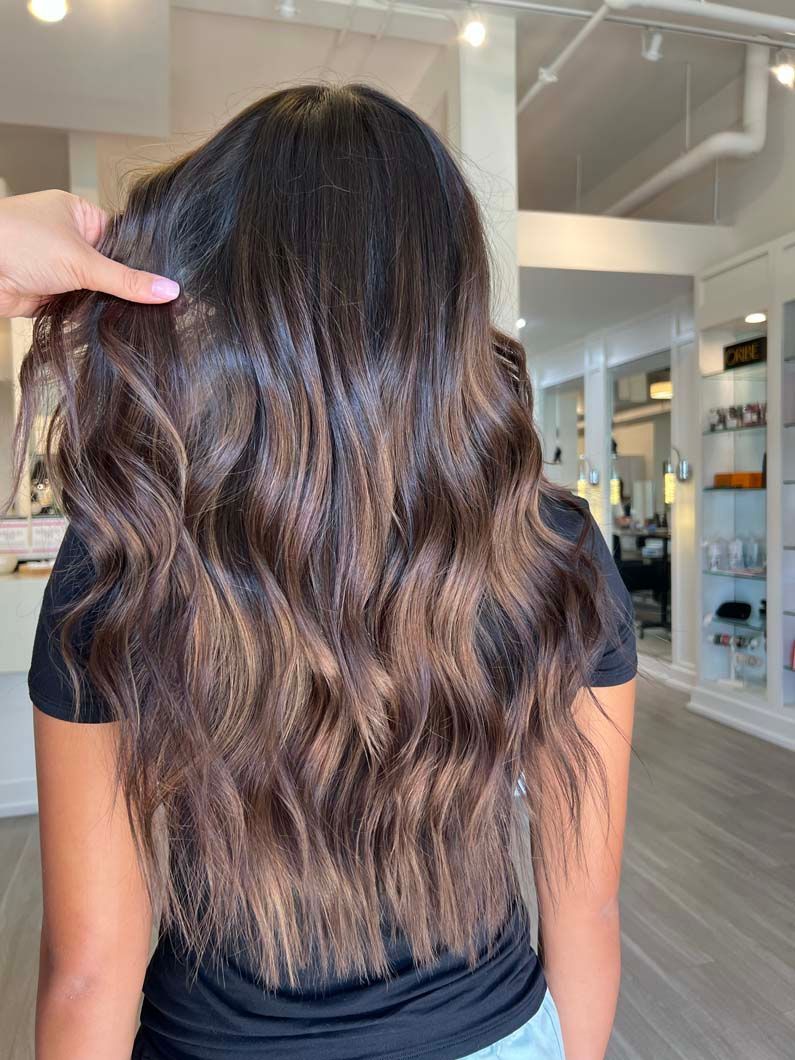 Woman's wavy, dark brown hair with brown balayage. A hand is holding a strand of hair inside a salon.