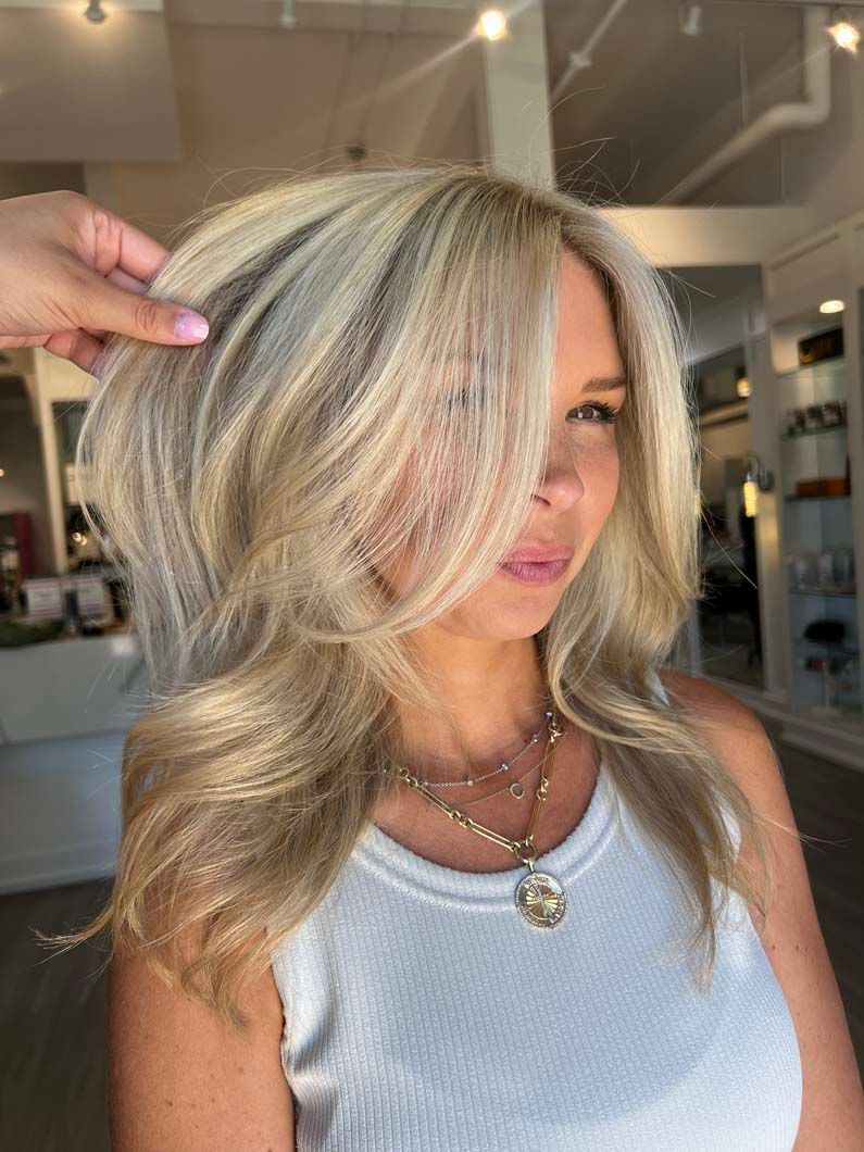 Woman with blonde hair styled, hand fluffing it, wearing a white tank top, in a salon.