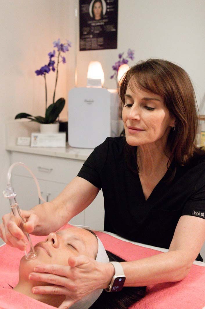 Woman performing a facial treatment on a client, using a device; treatment room setting.