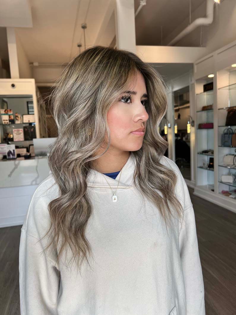 Woman with wavy, light brown hair wearing a cream sweatshirt, standing in a salon.