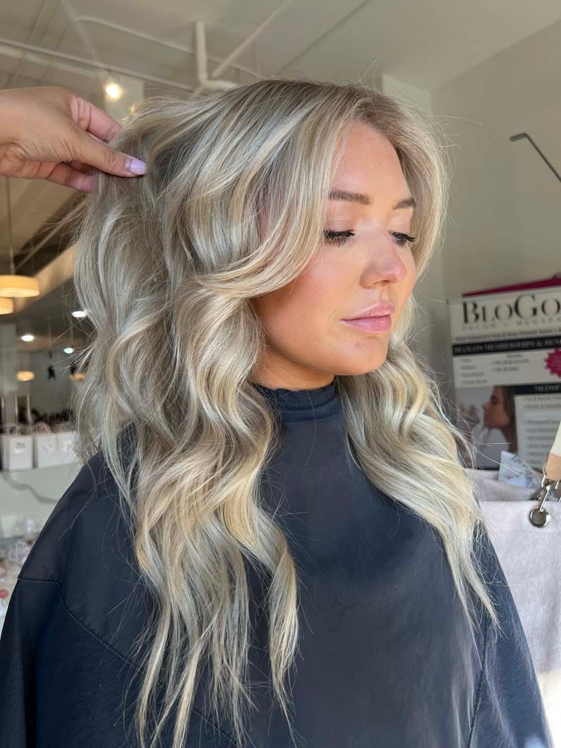 Woman with wavy, blonde hair at a salon. A hand adjusts her hair. She is wearing a black cape.