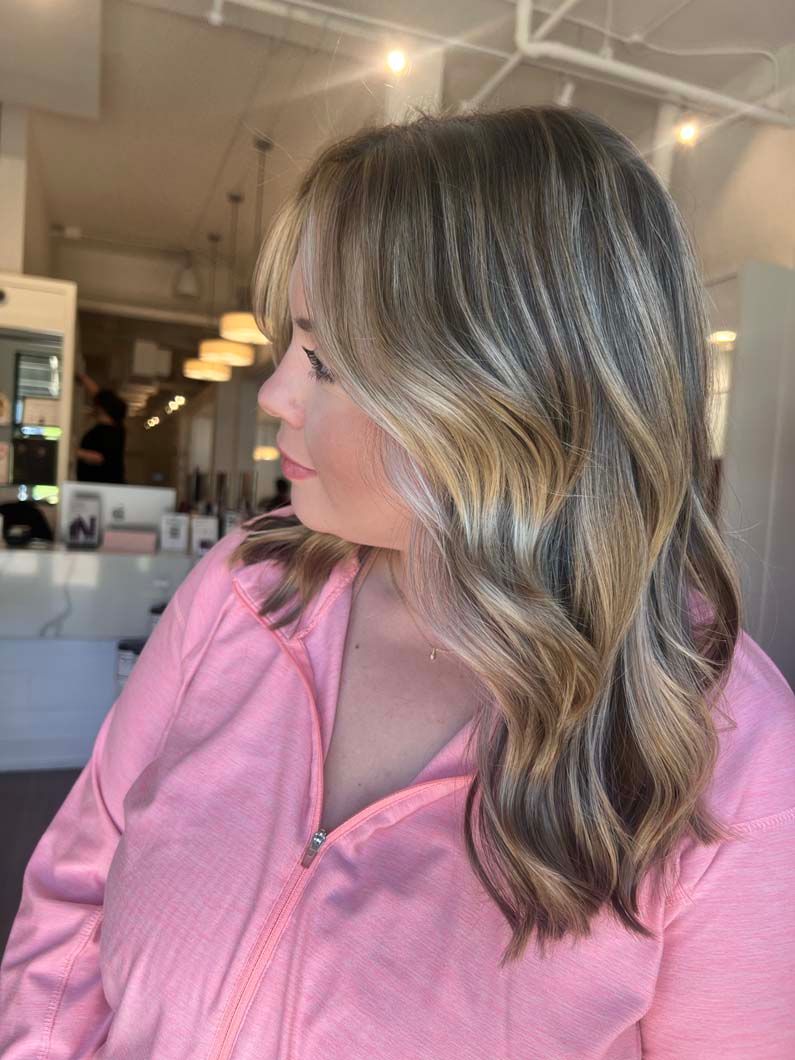Woman with wavy, highlighted hair wearing a pink jacket, in a salon setting.