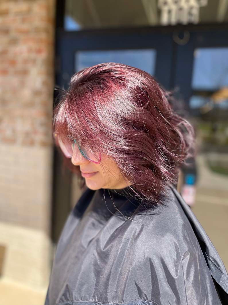 Woman with dark reddish-purple hair styled in loose waves, wearing a salon cape; outdoors.