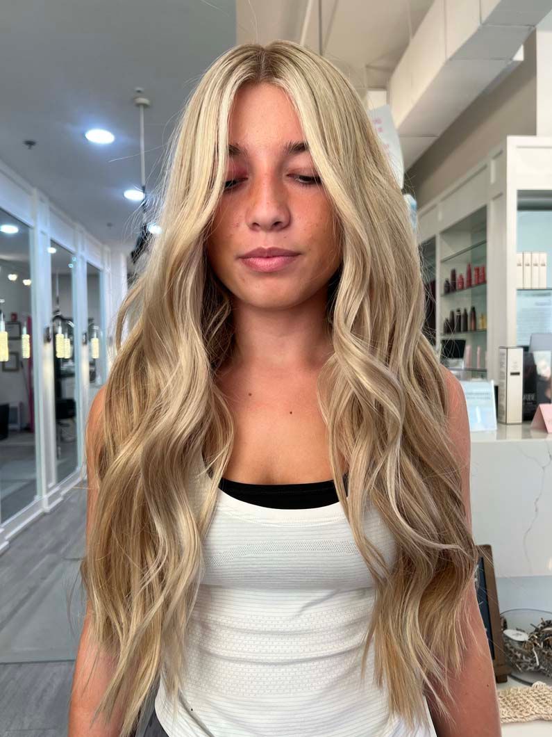Woman with wavy blonde and light brown hair in a salon, wearing a white tank top.