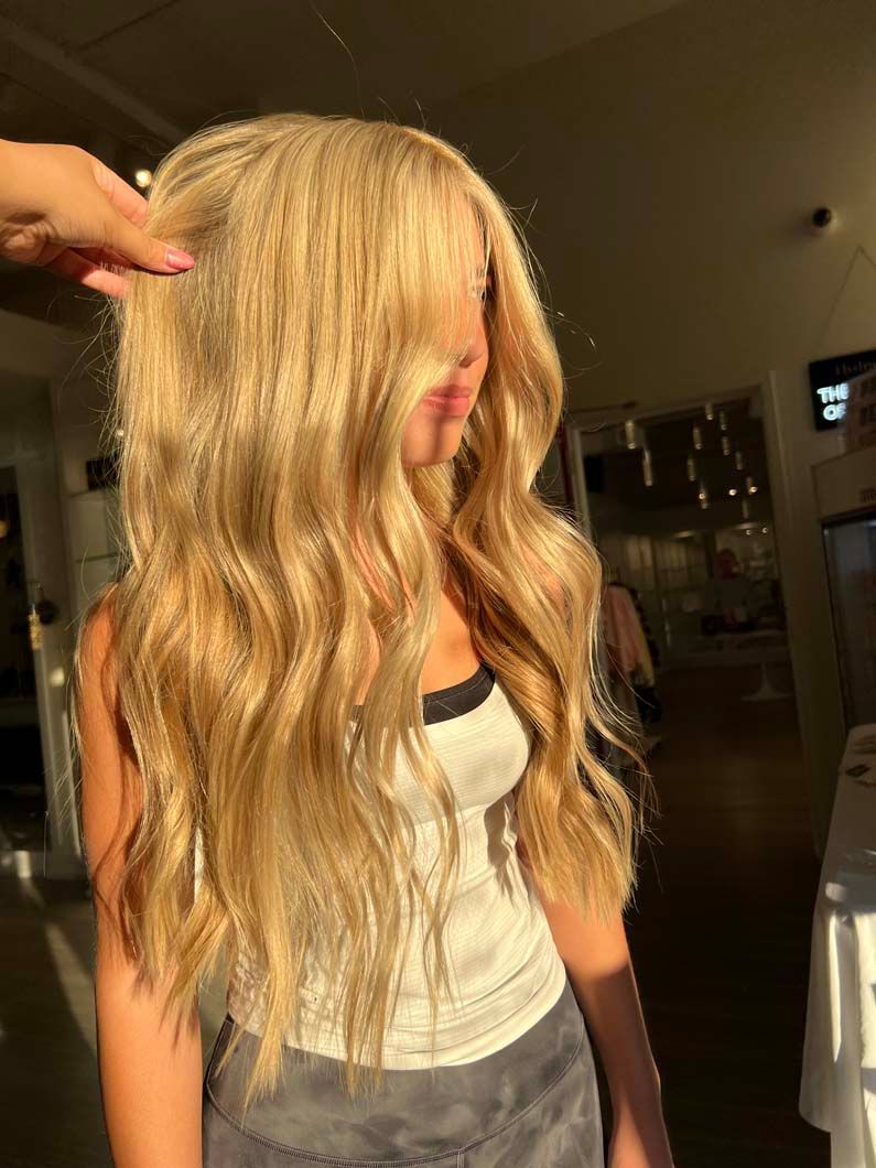 Woman with long, wavy blonde hair; wearing a white tank top and gray pants.