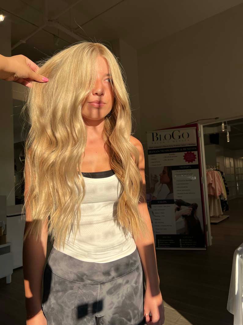 Woman with long blonde wavy hair, wearing a white tank top and grey pants, in an indoor setting with natural light.