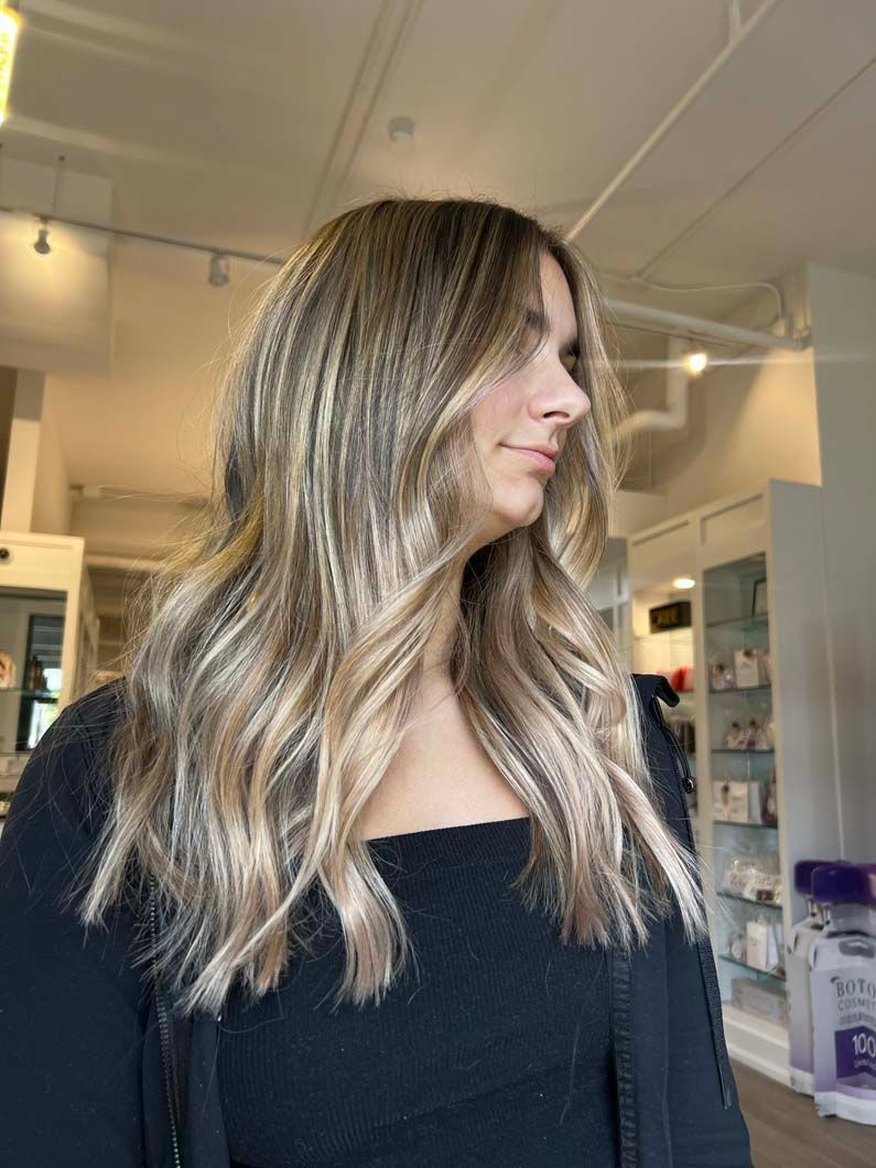 Woman with long, wavy blonde and brown hair in a salon, wearing a black top.