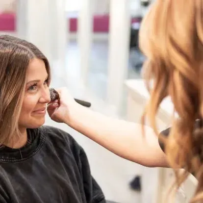Woman getting makeup applied with a brush by another person in a salon.