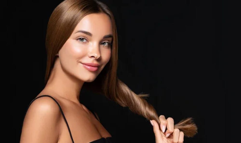 Woman with smooth, light brown hair, smiles, holding a section of her hair against a black background.