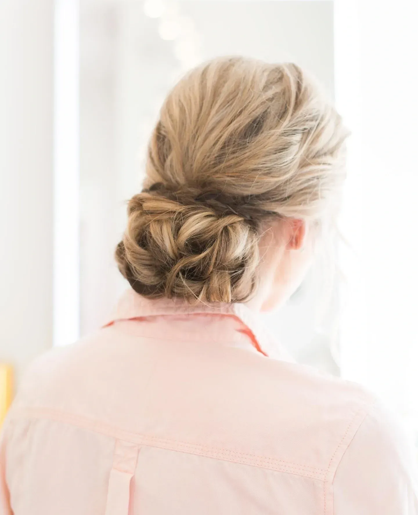 Woman with light brown hair in an updo, wearing a pink shirt, standing in front of a white background.