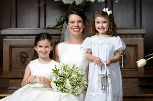 Bride and two flower girls smiling in front of a church altar. The bride holds a bouquet, and the flower girls hold a basket and lantern.