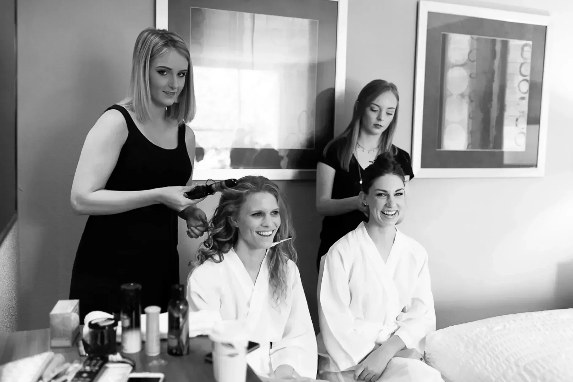 Two women in robes getting their hair styled by two other women in a room; makeup supplies on a table.