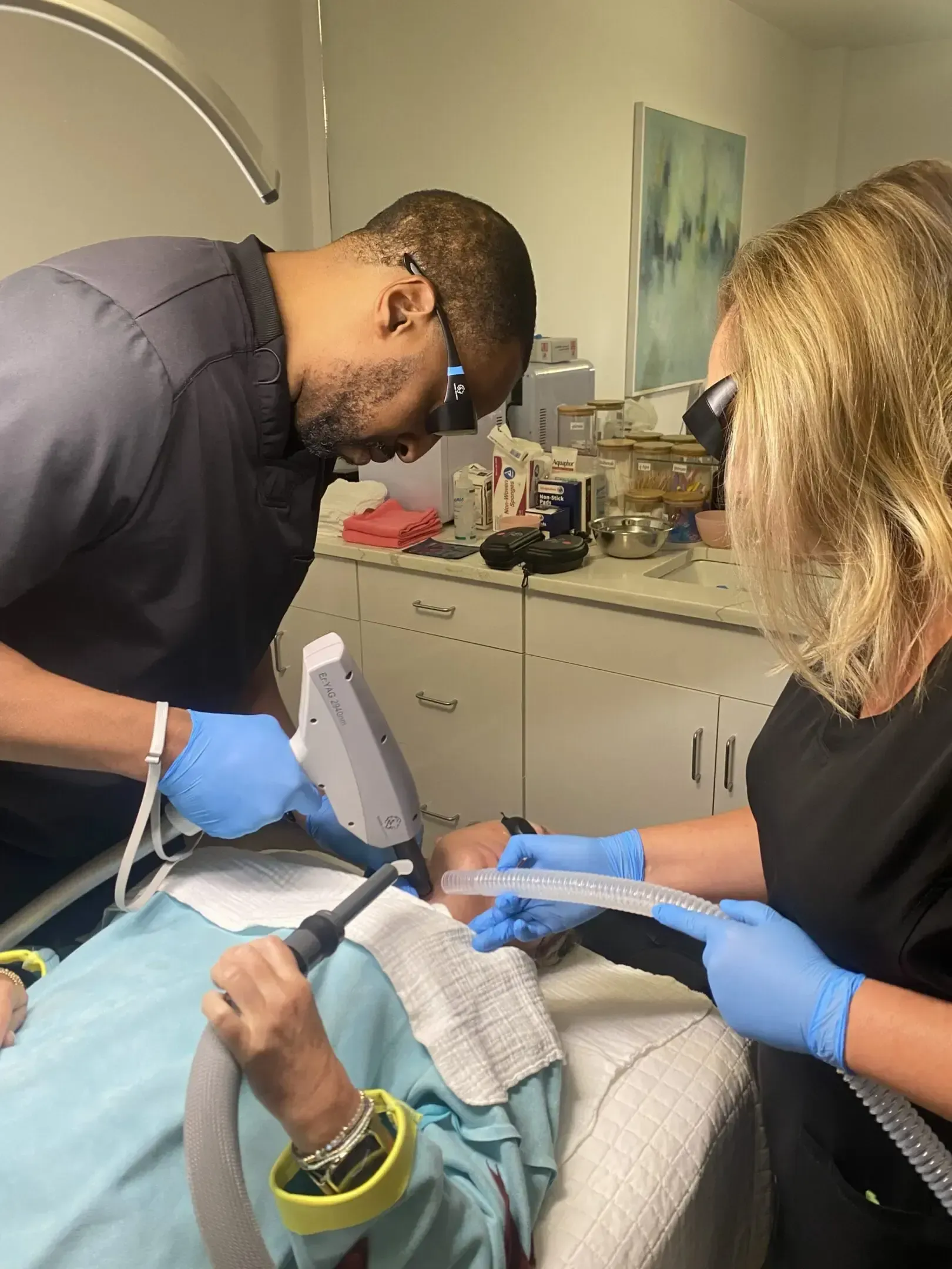 Two people using equipment on a patient in a medical setting, likely a dermatology procedure.