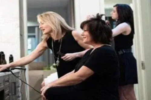 Three women in a salon: Stylist styling a woman's hair while another woman works in the background.