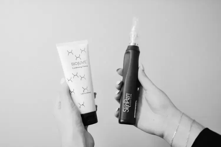 Person holding a tube of skincare cream and a microneedling pen in black and white.