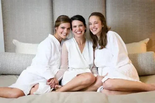 Three women in white robes smiling on a bed.