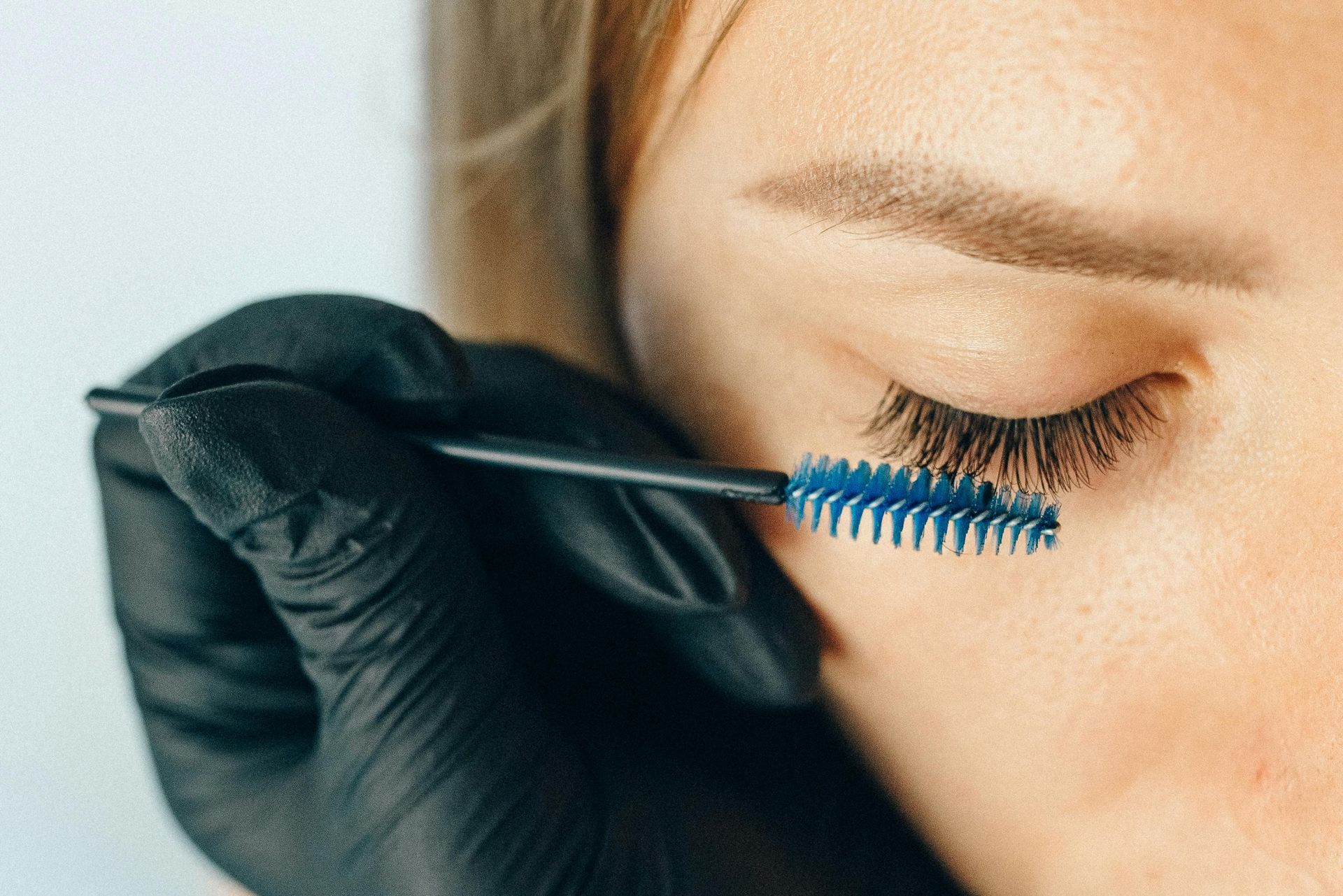 A person's closed eye with long eyelashes being brushed with a blue eyelash comb.