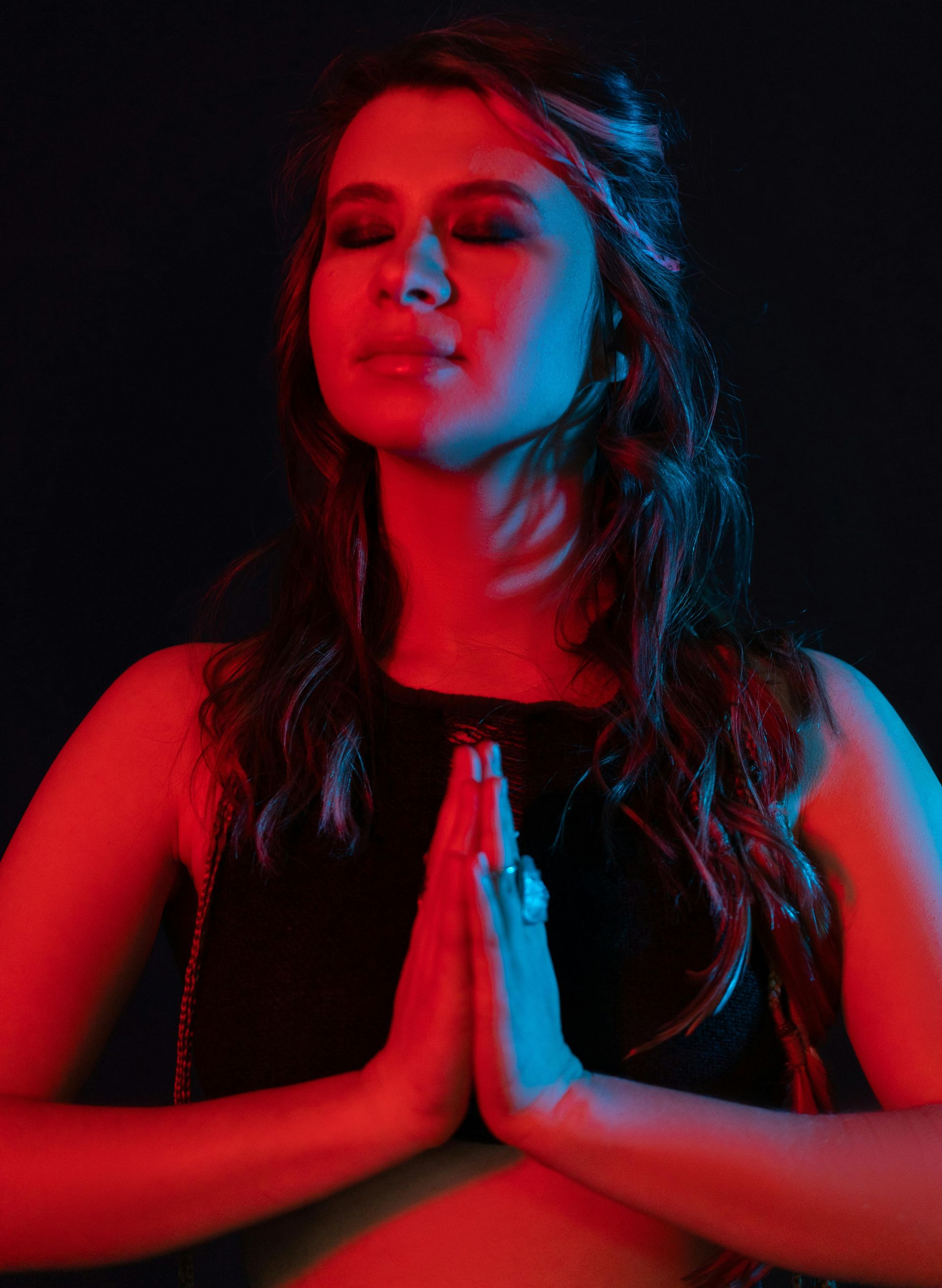 Woman with eyes closed, hands in prayer pose, lit with red and blue light, dark background.