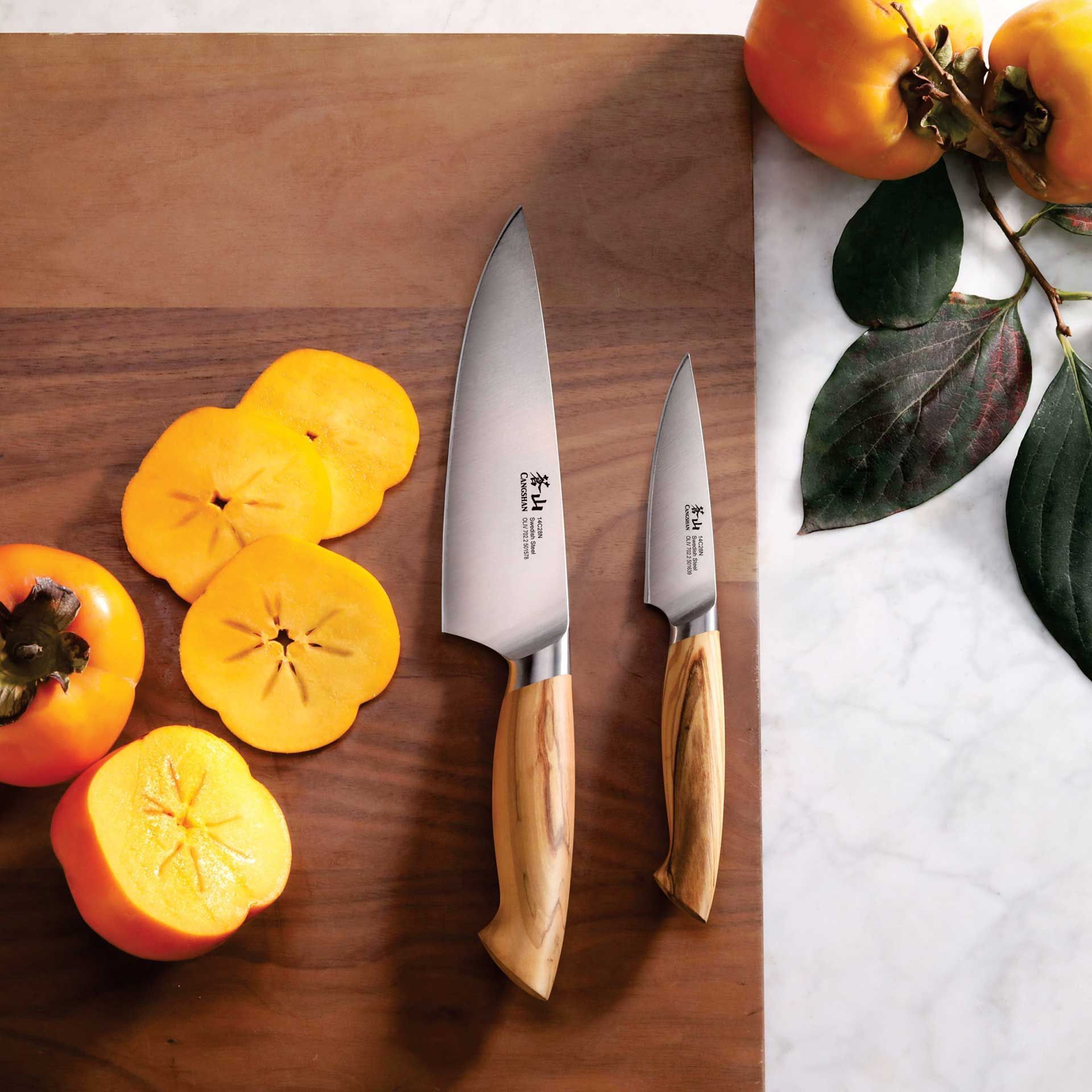 Two knives are on a wooden cutting board next to sliced oranges