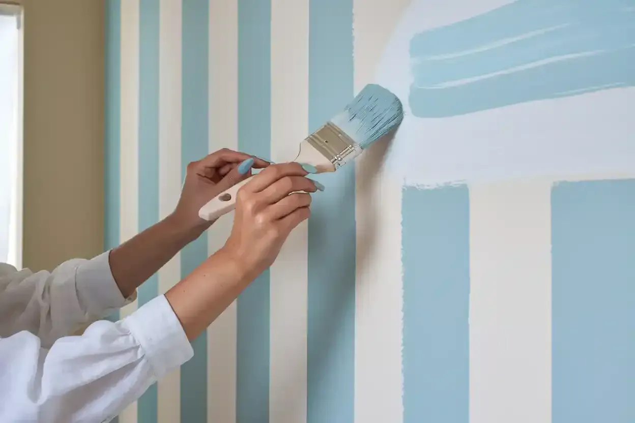 Hands painting a blue stripe on a wall with vertical blue and white stripes, using a paintbrush.