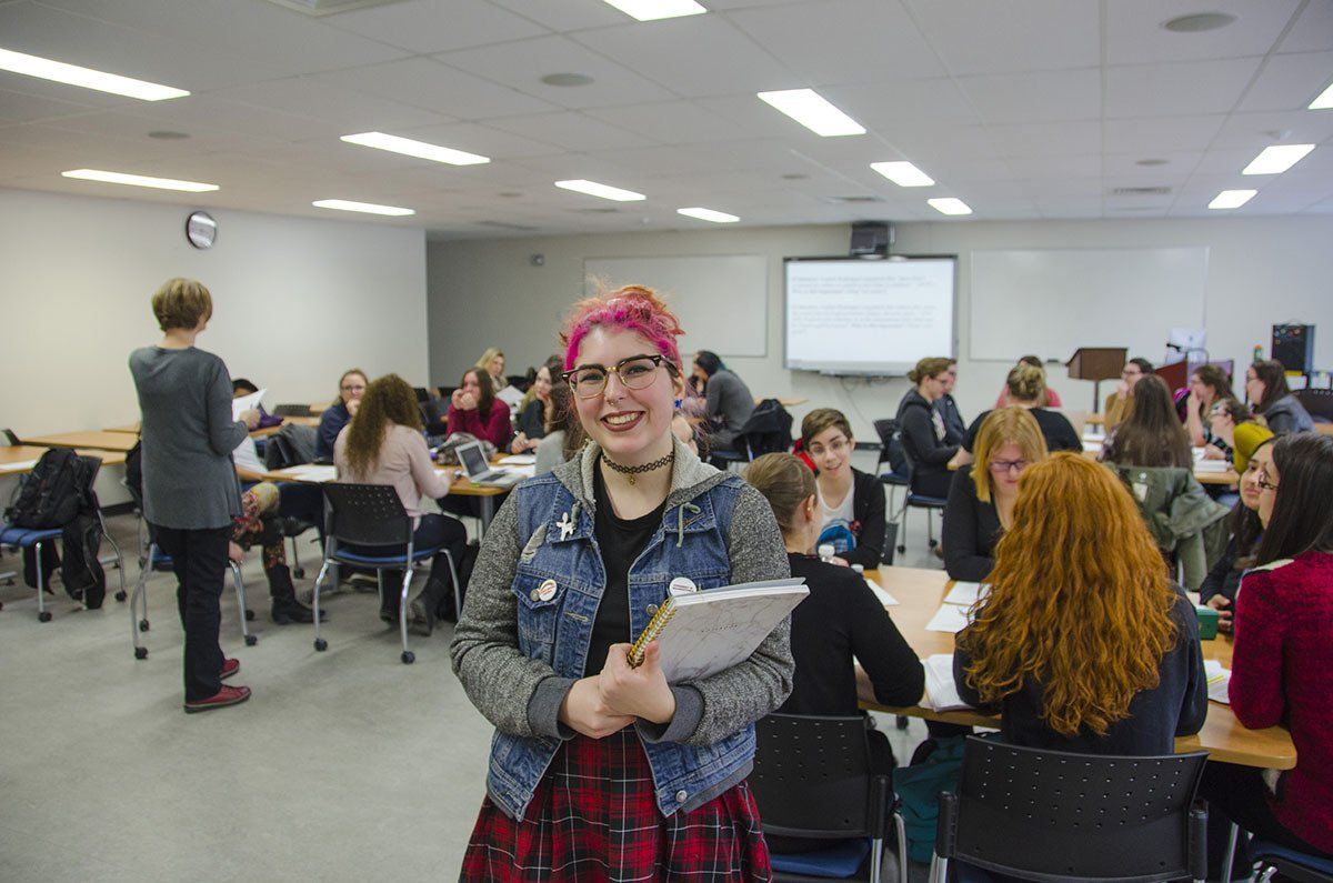 A woman with pink hair is standing in a classroom holding a clipboard.