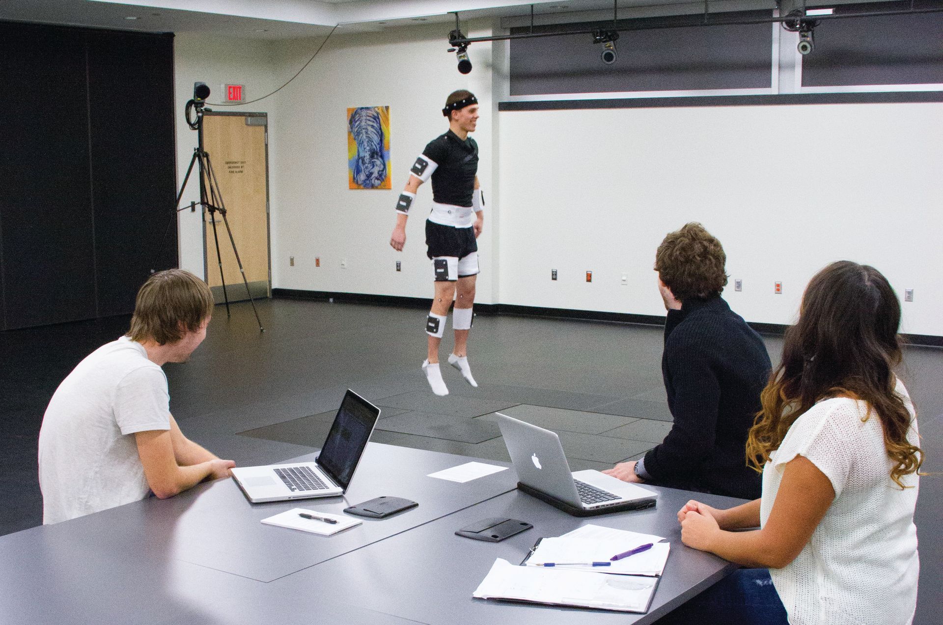 A group of people are sitting at a table with laptops in front of a man jumping in the air.
