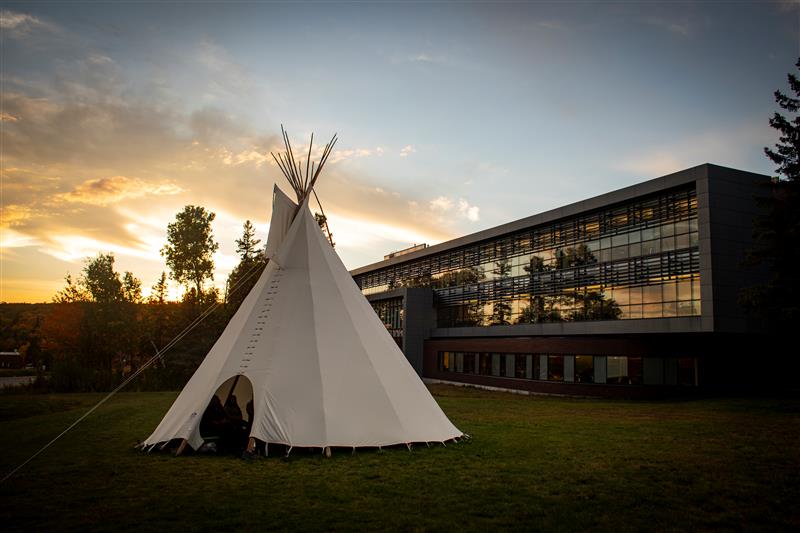 A teepee is sitting in the grass in front of a building at sunset.