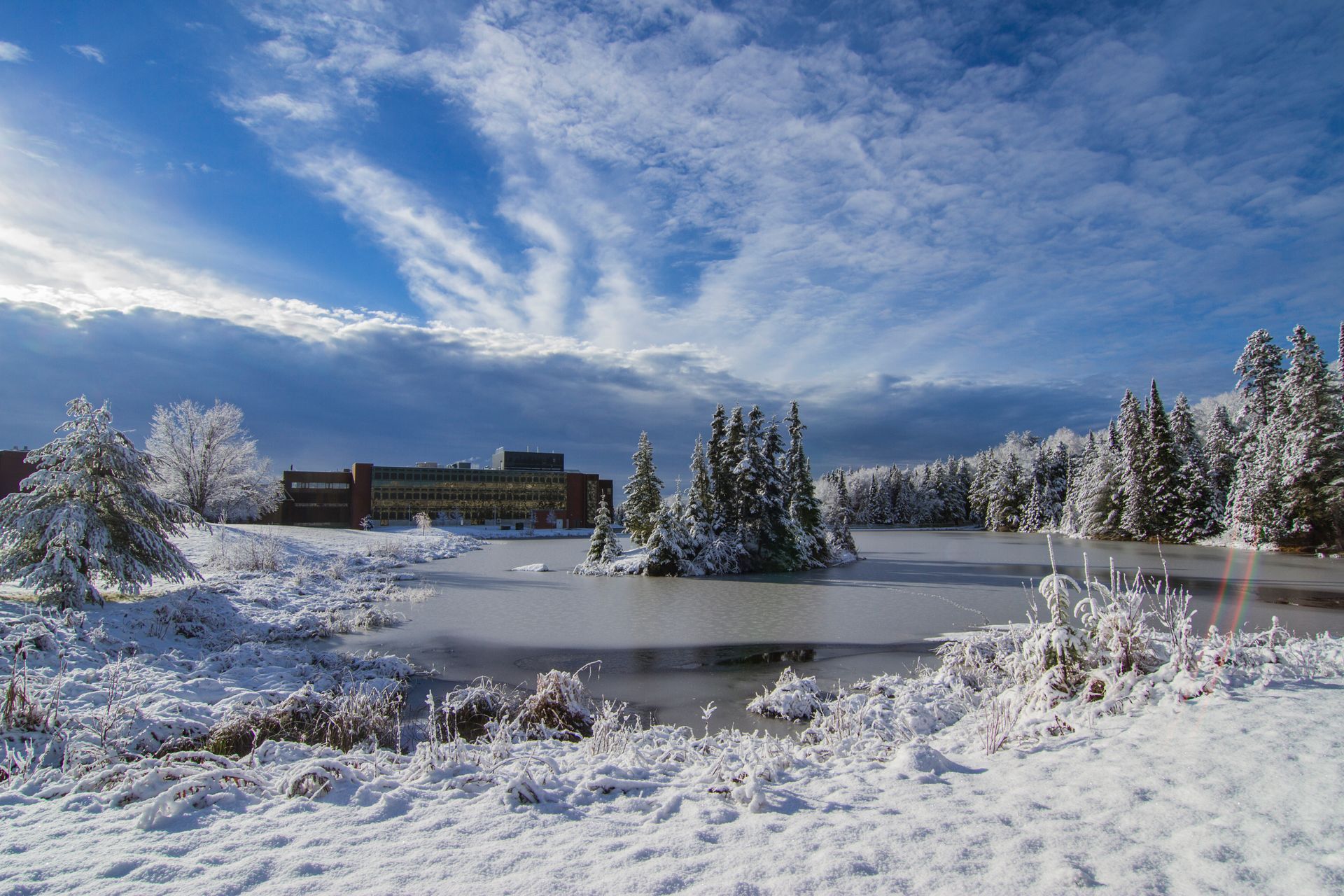 A snowy landscape with a lake and trees in the foreground and a building in the background.