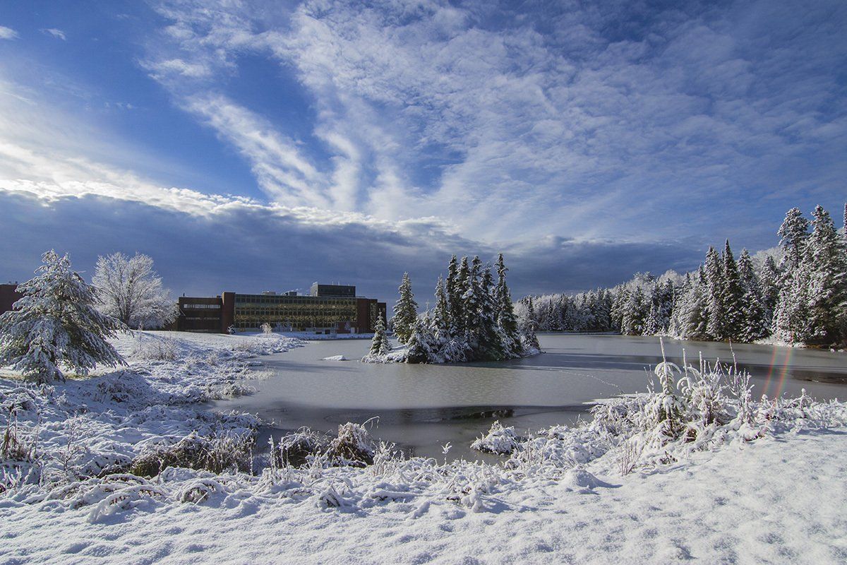 A frozen lake surrounded by snow covered trees with a building in the background.