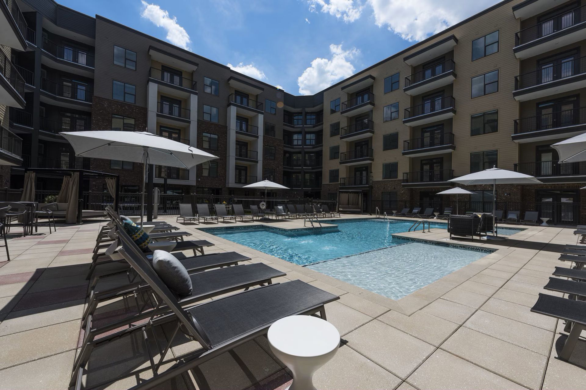 Pool area with lounge chairs, umbrellas, and a modern apartment building. Sunny day.
