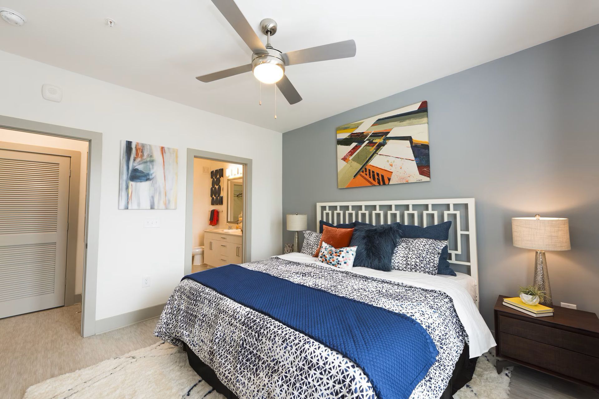Bedroom with a patterned bedspread, blue and orange pillows, and colorful artwork on a gray accent wall.