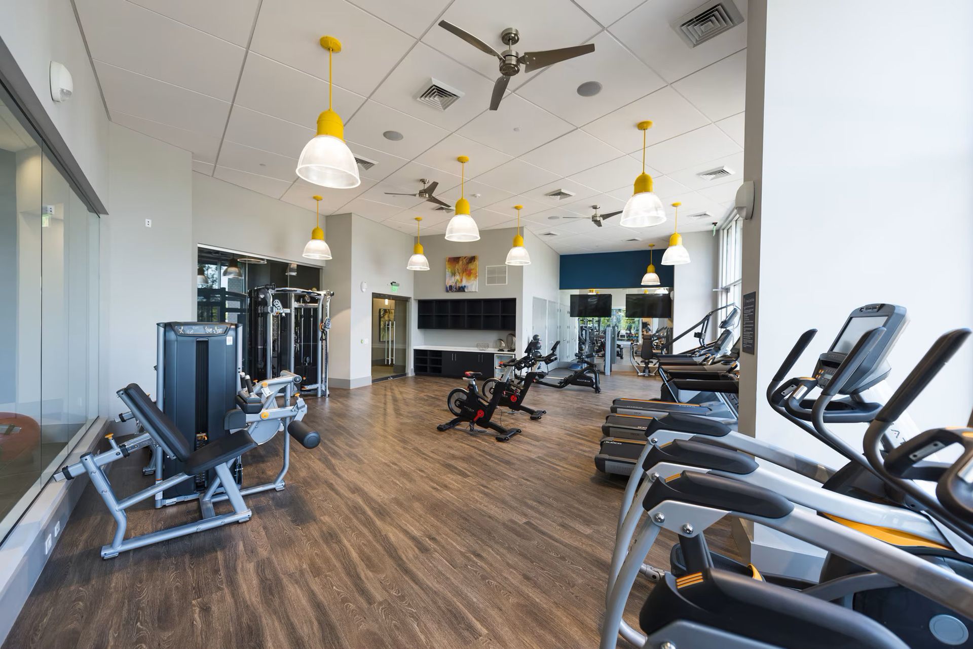 Gym interior with fitness equipment, wooden floor, yellow pendant lights, and a ceiling fan.
