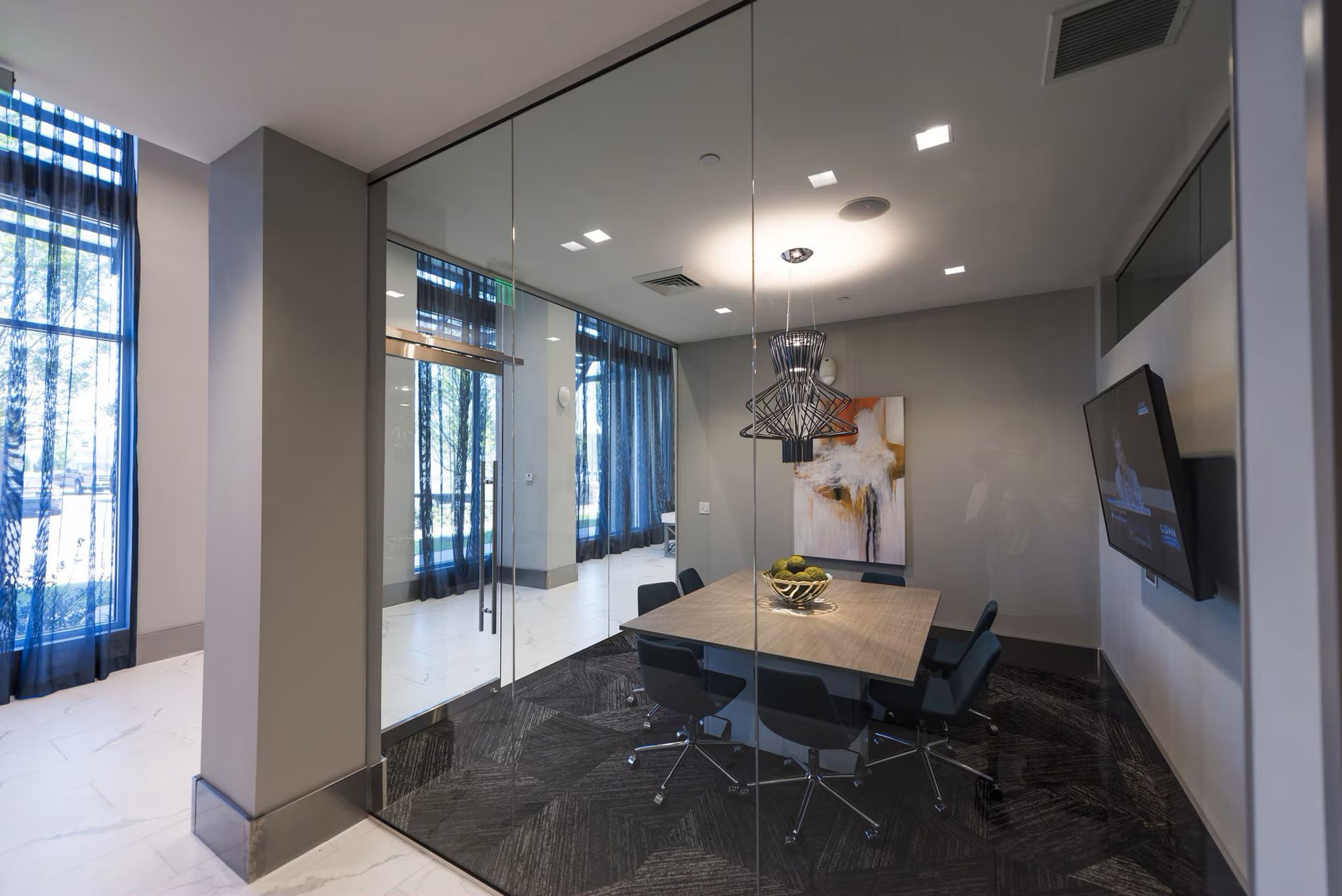 Glass-walled meeting room with a table, chairs, and a painting, inside a building, with a dark floor.