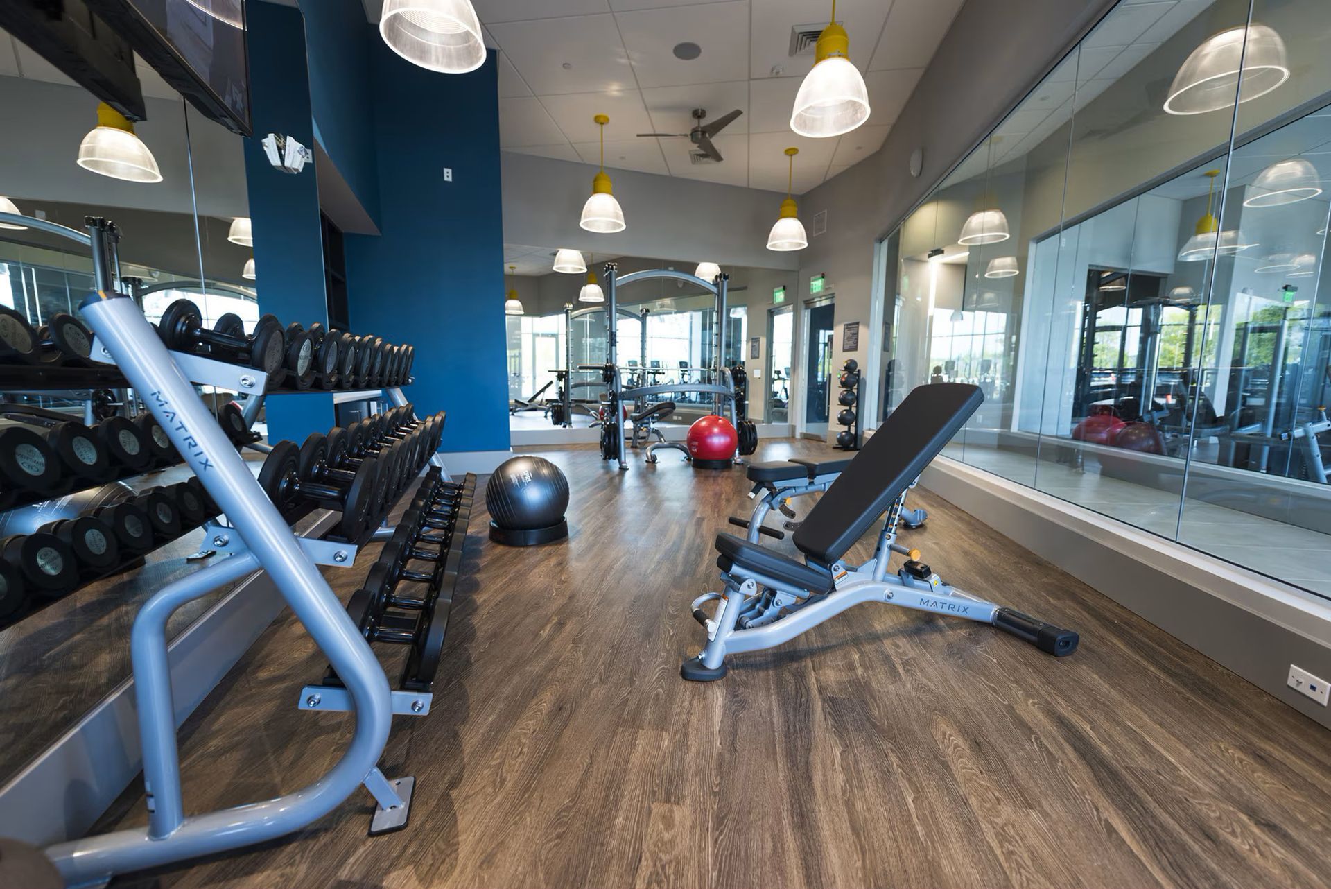 Gym with dumbbells, bench, and exercise equipment, reflective mirrors, and wooden floors.